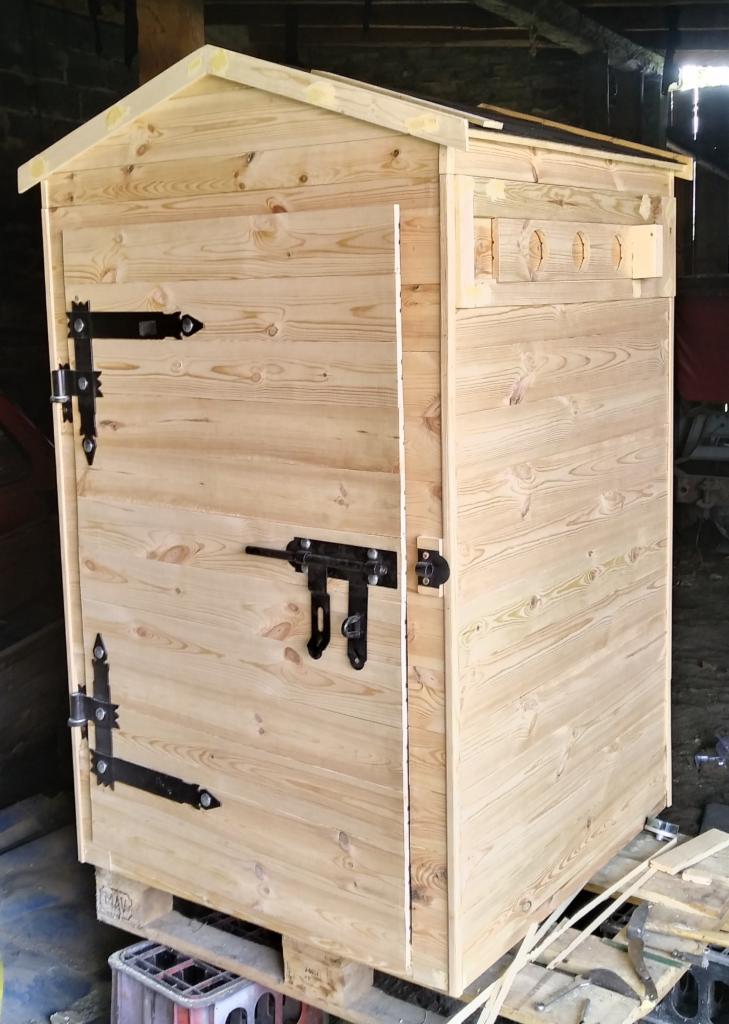 Surowa, drewniana wędzarnia z czarnymi okuciami, umieszczona na palecie, widoczne otwory wentylacyjne i proste zapięcie, tło warsztatowe.