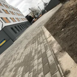 Ułożona, szara kostka brukowa na chodniku przed budynkiem z logo firmy Maxbud, widoczne nierówności terenu po prawej stronie.