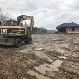 Żółta koparka CAT M312 na błotnistym placu budowy z widocznym w tle niedokończonym domem z czerwonej cegły pod czarnym dachem.