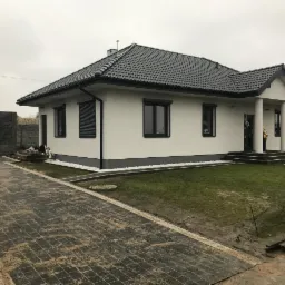 Nowoczesny dom jednorodzinny z ciemną dachówką i jasną elewacją, widoczny z perspektywy świeżo ułożonej kostki brukowej na podjeździe, fragment trawnika i dekoracyjne kamienie wzdłuż fundamentów.