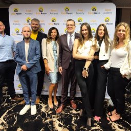 Greathouse - Grupa osób pozuje na tle baneru reklamowego GreatHouse Academy z logo domu, prawdopodobnie podczas wydarzenia firmowego, na marmurowej podłodze.