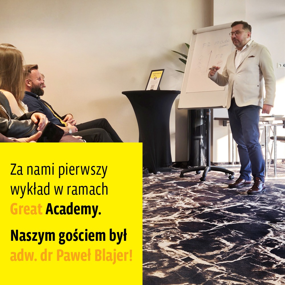 Prelegent w beżowej marynarce i okularach prowadzi wykład, pisząc na flipcharcie; w tle siedzą słuchacze i stoi czarny stolik z żółtą tabliczką informacyjną; na podłodze wzorzysta wykładzina.