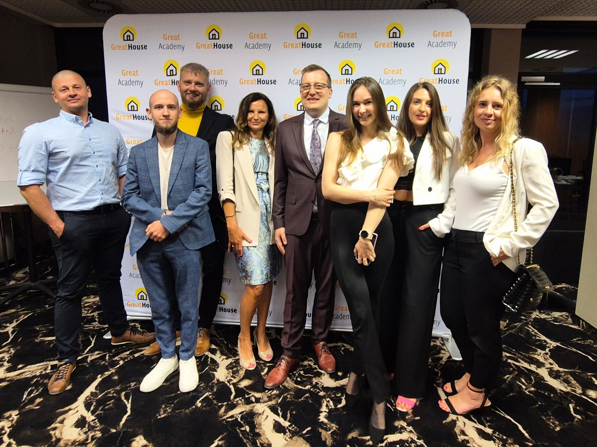 Grupa osób pozuje na tle baneru reklamowego GreatHouse Academy z logo domu, prawdopodobnie podczas wydarzenia firmowego, na marmurowej podłodze.