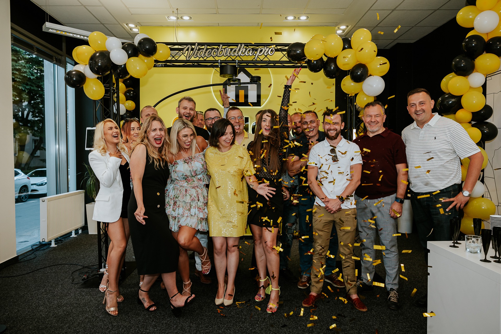 Grupa uśmiechniętych osób świętujących pod logo Videobudka.pro, z konfetti, balonami w kolorach żółtym, białym i czarnym, w jasnym wnętrzu z oknami.