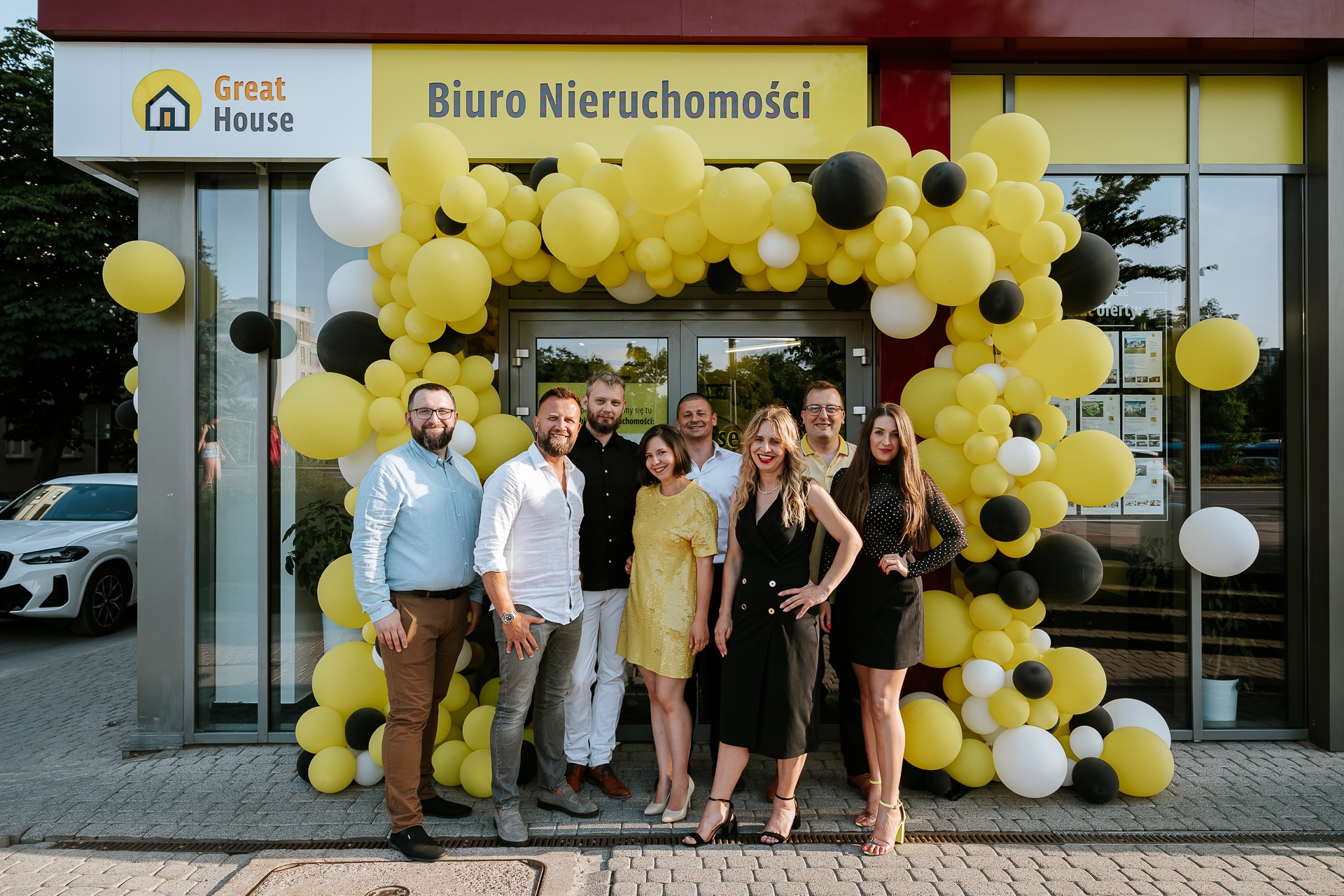 Grupa pracowników biura nieruchomości 'Great House' pozuje przed wejściem udekorowanym żółto-biało-czarnymi balonami.
