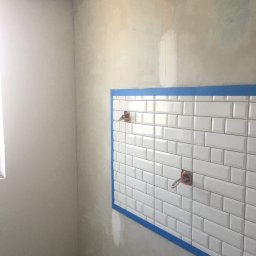 Marcin Lewandowski - Fragment łazienki w trakcie prac wykończeniowych: białe, prostokątne płytki ułożone na ścianie, zabezpieczone niebieską taśmą malarską; widoczne wystające kable i rury instalacyjne, surowe ściany...