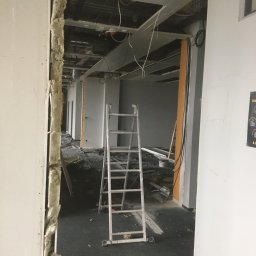 Marcin Lewandowski - Korytarz w trakcie prac wykończeniowych z odsłoniętą izolacją ściany, aluminiowa drabina w centrum kadru, widoczne kable i elementy instalacji pod sufitem.