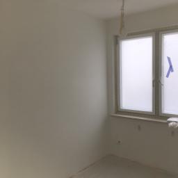 Marcin Lewandowski - Świeżo pomalowane na biało wnętrze pokoju z oknem zabezpieczonym folią i taśmą malarską, podłoga również zabezpieczona folią, widoczne przewody elektryczne wystające z sufitu.