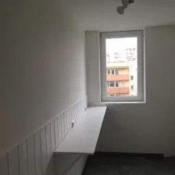 Wnętrze pokoju po remoncie: pomalowane ściany z dekoracyjną boazerią, zamontowany składany blat pod oknem, szara podłoga z płytek.