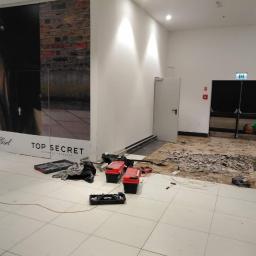 Marcin Lewandowski - Pracownik w zielonej koszulce usuwa starą podłogę w lokalu handlowym, widoczne narzędzia, gruz i częściowo zerwana nawierzchnia, w tle plakat reklamowy.