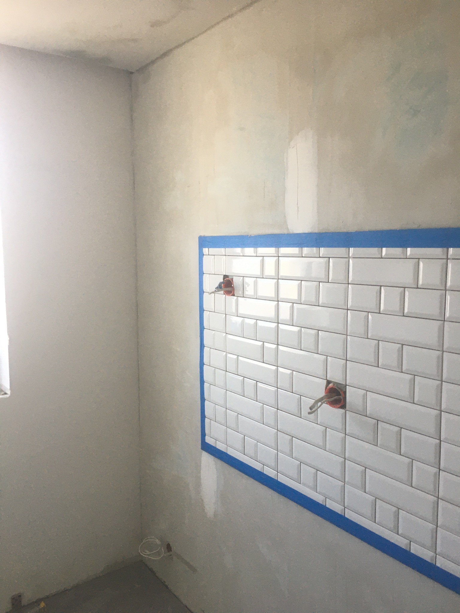 Fragment łazienki w trakcie prac wykończeniowych: białe, prostokątne płytki ułożone na ścianie, zabezpieczone niebieską taśmą malarską; widoczne wystające kable i rury instalacyjne, surowe ściany...