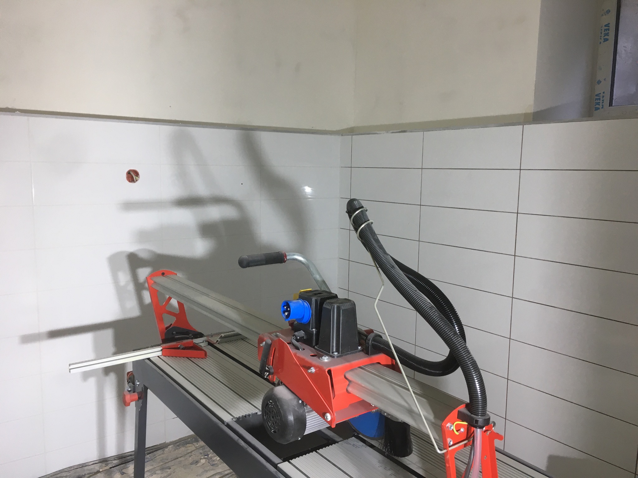 Elektryczna przecinarka do płytek ustawiona w rogu pomieszczenia z częściowo wyłożonymi białymi płytkami, widoczna instalacja elektryczna w ścianie.