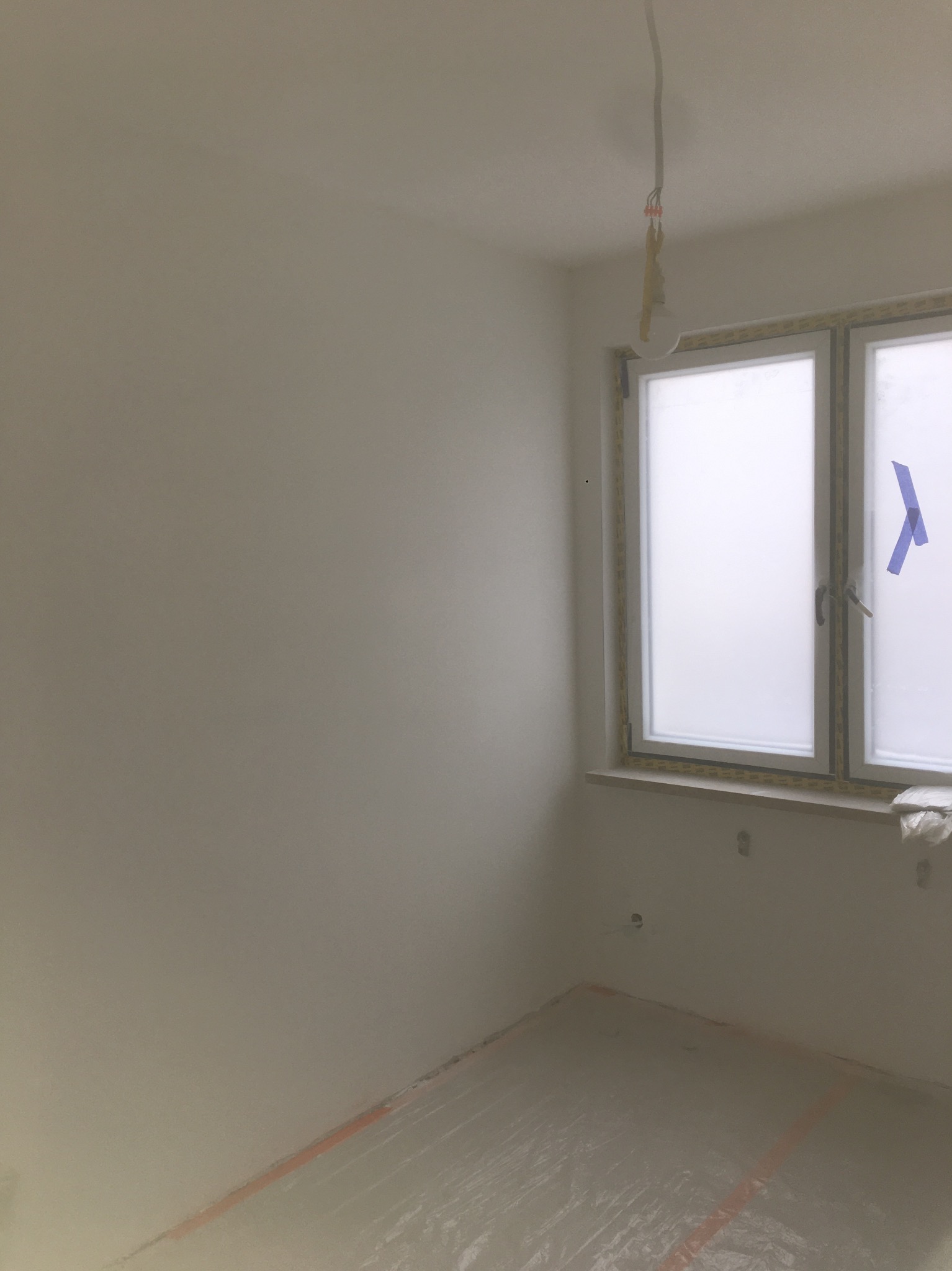 Świeżo pomalowane na biało wnętrze pokoju z oknem zabezpieczonym folią i taśmą malarską, podłoga również zabezpieczona folią, widoczne przewody elektryczne wystające z sufitu.