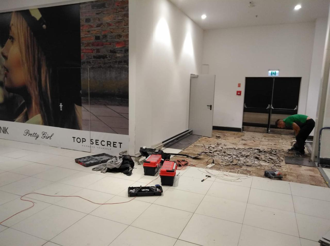 Pracownik w zielonej koszulce usuwa starą podłogę w lokalu handlowym, widoczne narzędzia, gruz i częściowo zerwana nawierzchnia, w tle plakat reklamowy.