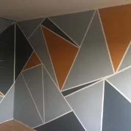 Geometryczny wzór na ścianie w odcieniach szarości i pomarańczu, wyznaczony za pomocą białej taśmy malarskiej, widoczny fragment podłogi.