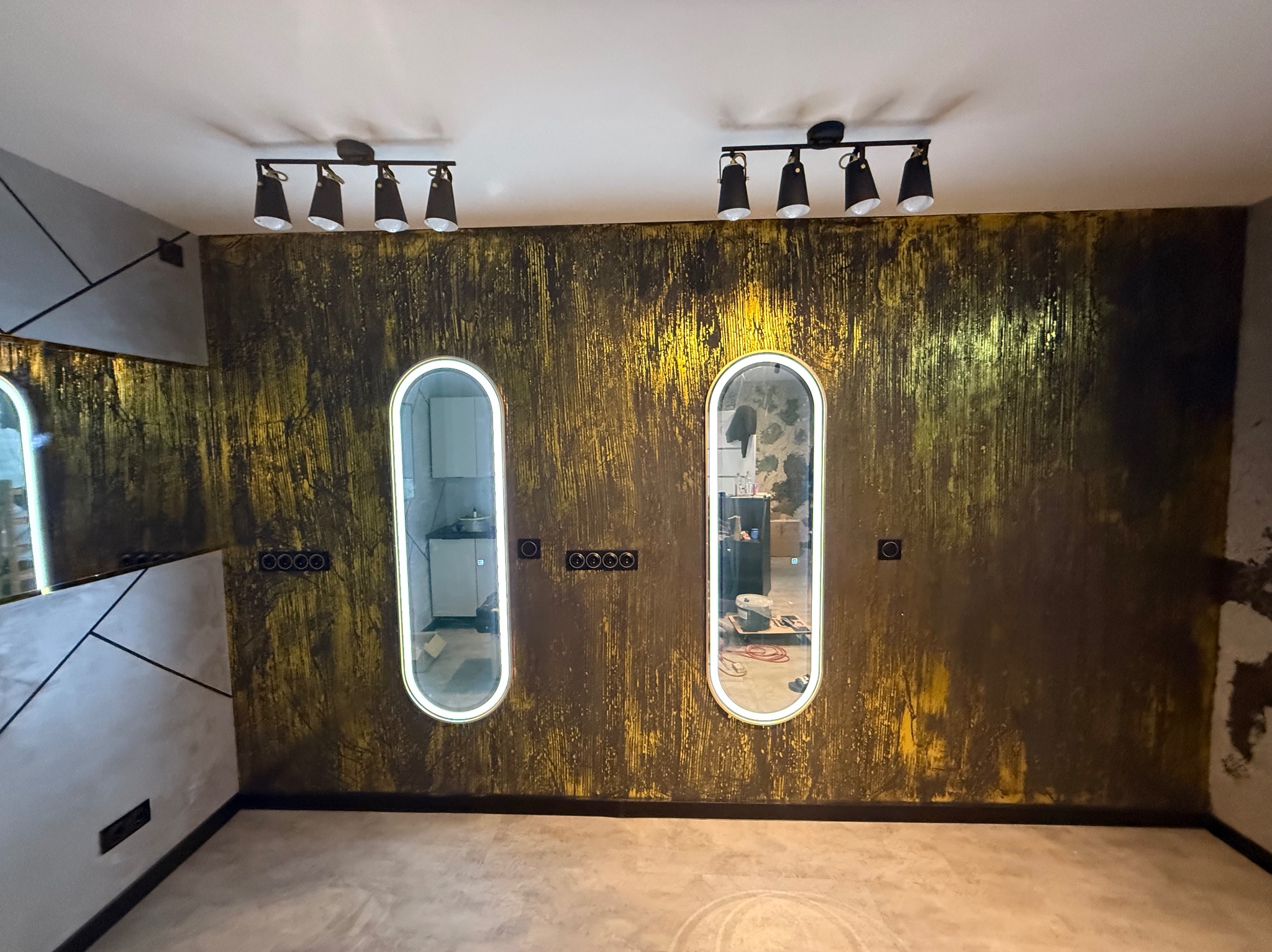Wnętrze po remoncie: ściana z efektem dekoracyjnym w odcieniach złota i czerni, dwa podświetlane, owalne lustra. Widoczne gniazdka, oświetlenie sufitowe i fragmenty mebli.