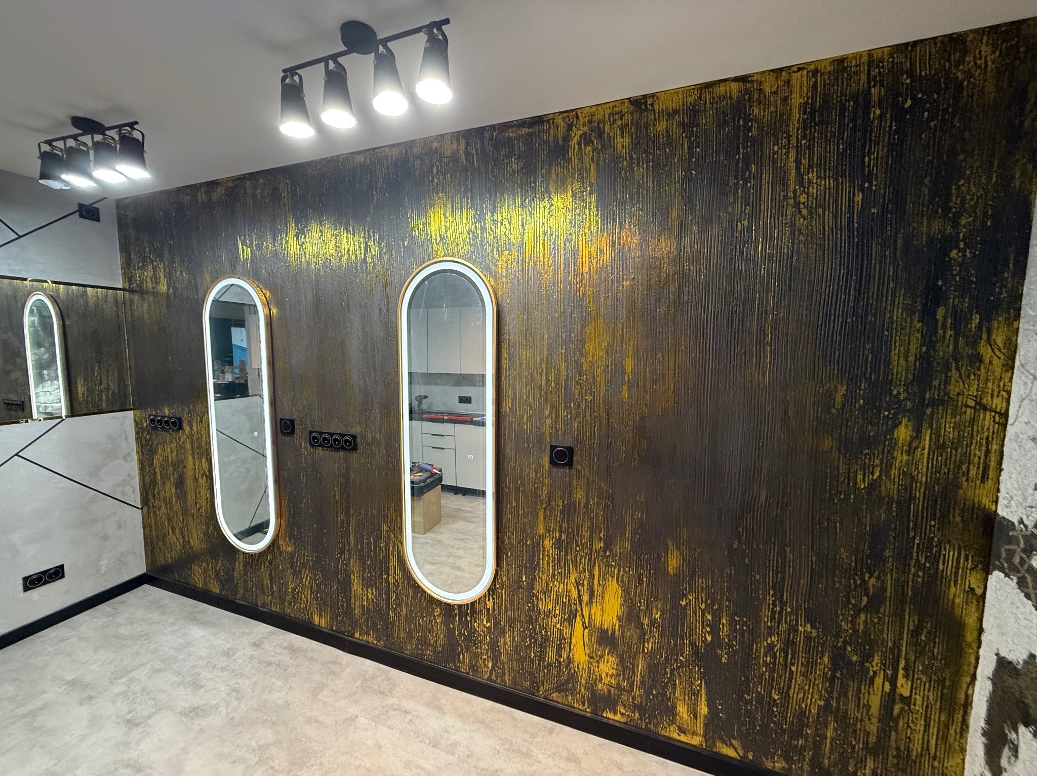 Ściana z dekoracyjną, ciemną fakturą i złotymi akcentami, podświetlane lustra w złotych ramach, czarne gniazdka, nowoczesne oświetlenie sufitowe.