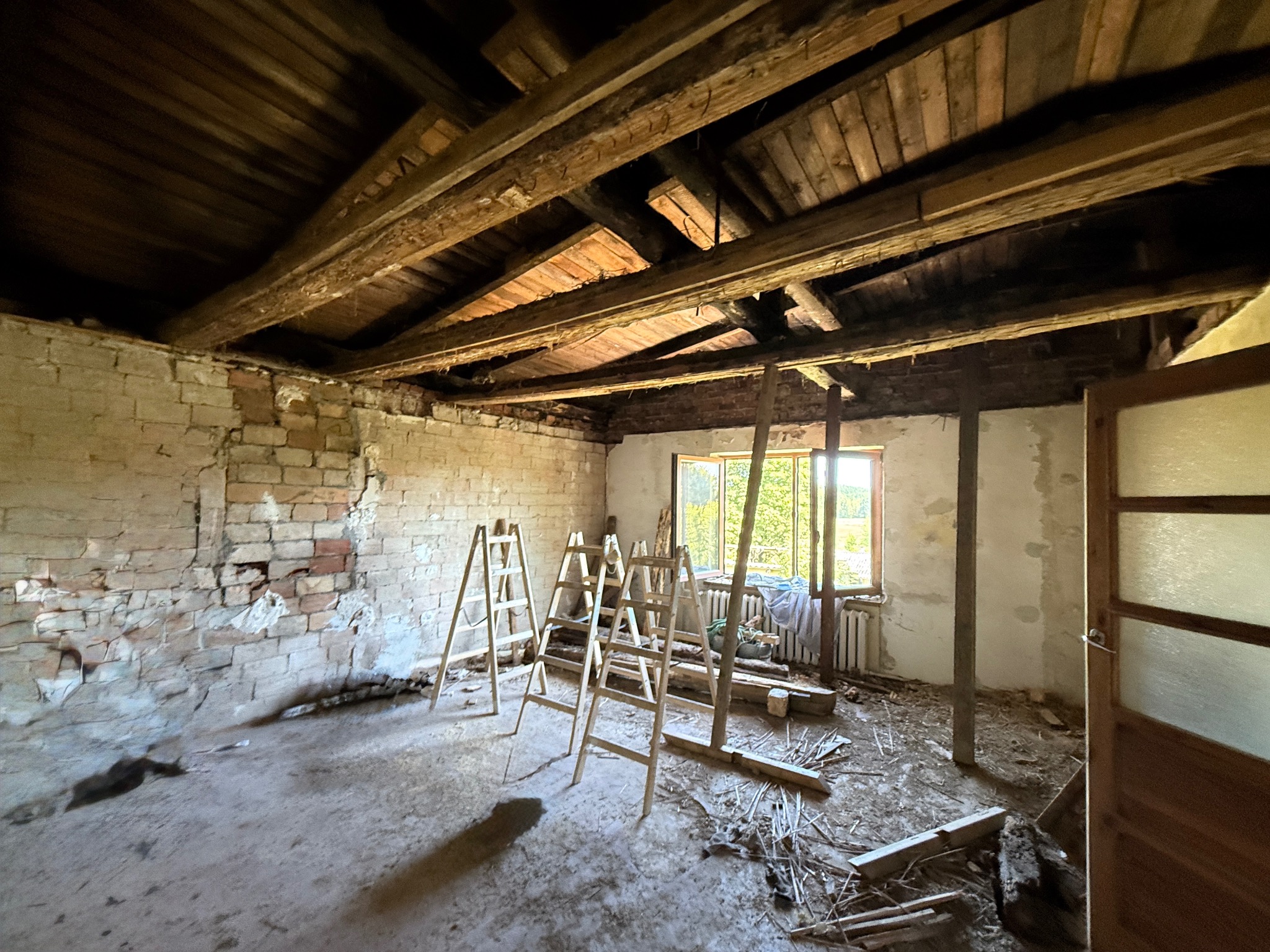 Wnętrze remontowanego pomieszczenia z odsłoniętą cegłą, drewnianym stropem i trzema drabinami. Widoczne ślady prac remontowych na podłodze i ścianach. Okno z widokiem na zieleń.