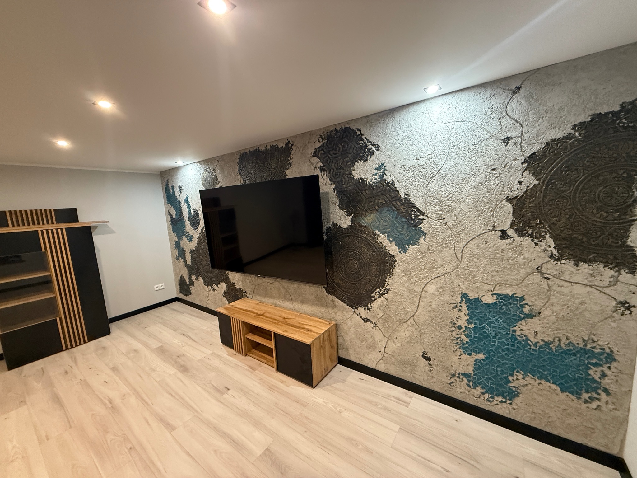 Efektowna ściana z dekoracyjnym tynkiem strukturalnym w odcieniach szarości, brązu i błękitu, oświetlona punktowo. Na ścianie wisi telewizor, a pod nim szafka RTV.