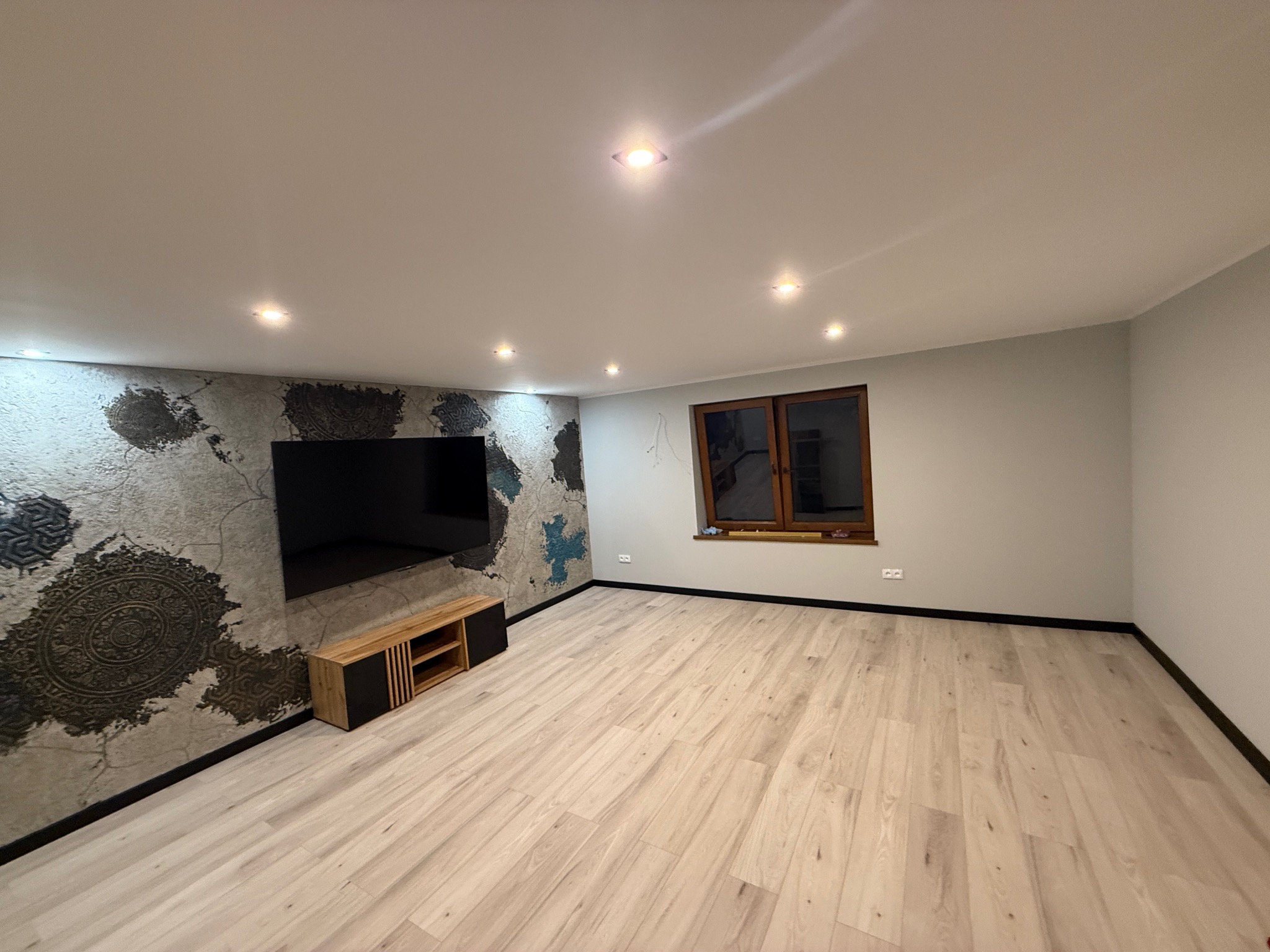 Wykończone wnętrze pokoju z podłogą z paneli, dekoracyjną ścianą z motywami mandali, oświetleniem punktowym i oknem z brązową ramą. Nowoczesny design.