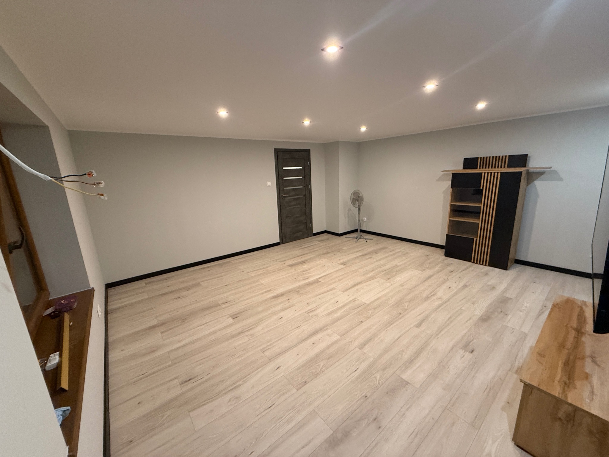 Jasne panele podłogowe w nowocześnie wykończonym pokoju z szarymi ścianami, czarnymi listwami przypodłogowymi i minimalistycznym regałem. Widoczne przewody elektryczne.