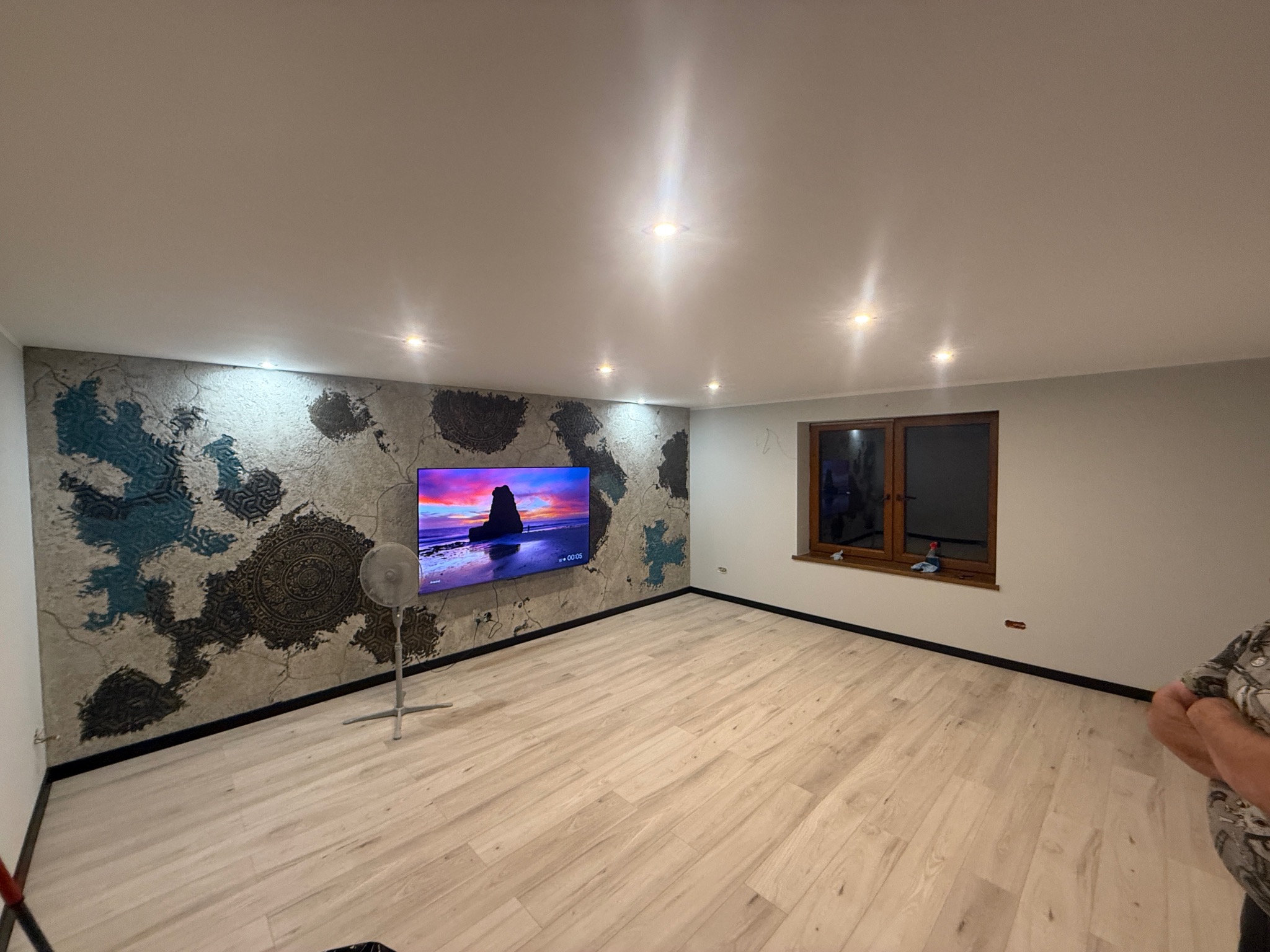 Wykończone wnętrze pokoju z panelami podłogowymi i ścianą z dekoracyjnym tynkiem imitującym mapę świata z motywami mandali. Na ścianie wisi telewizor.