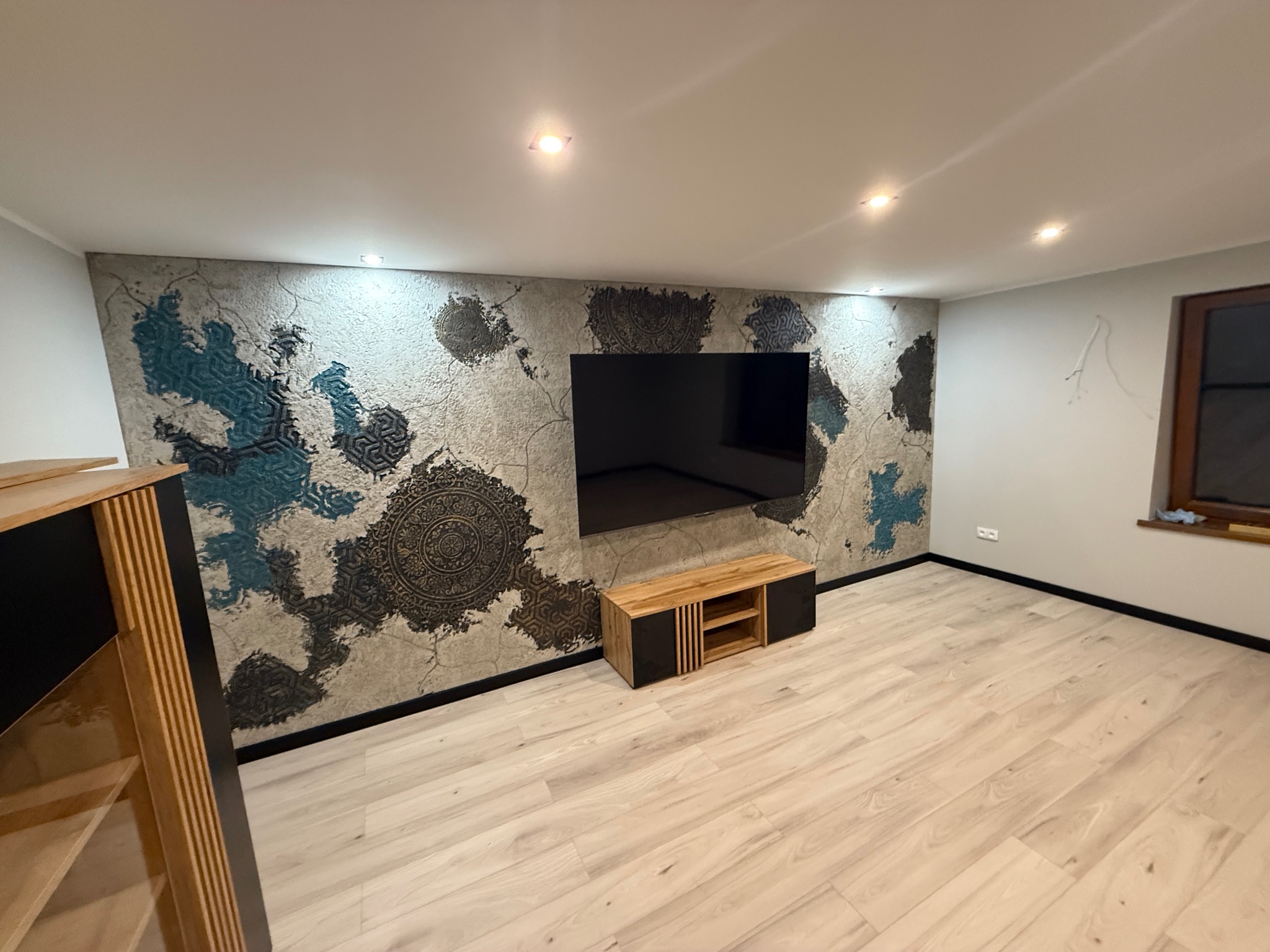 Wykończenie wnętrza: ściana z dekoracyjnym tynkiem w stylu etno, czarny telewizor, drewniane meble i jasna podłoga. Elementy niebieskie i złote.