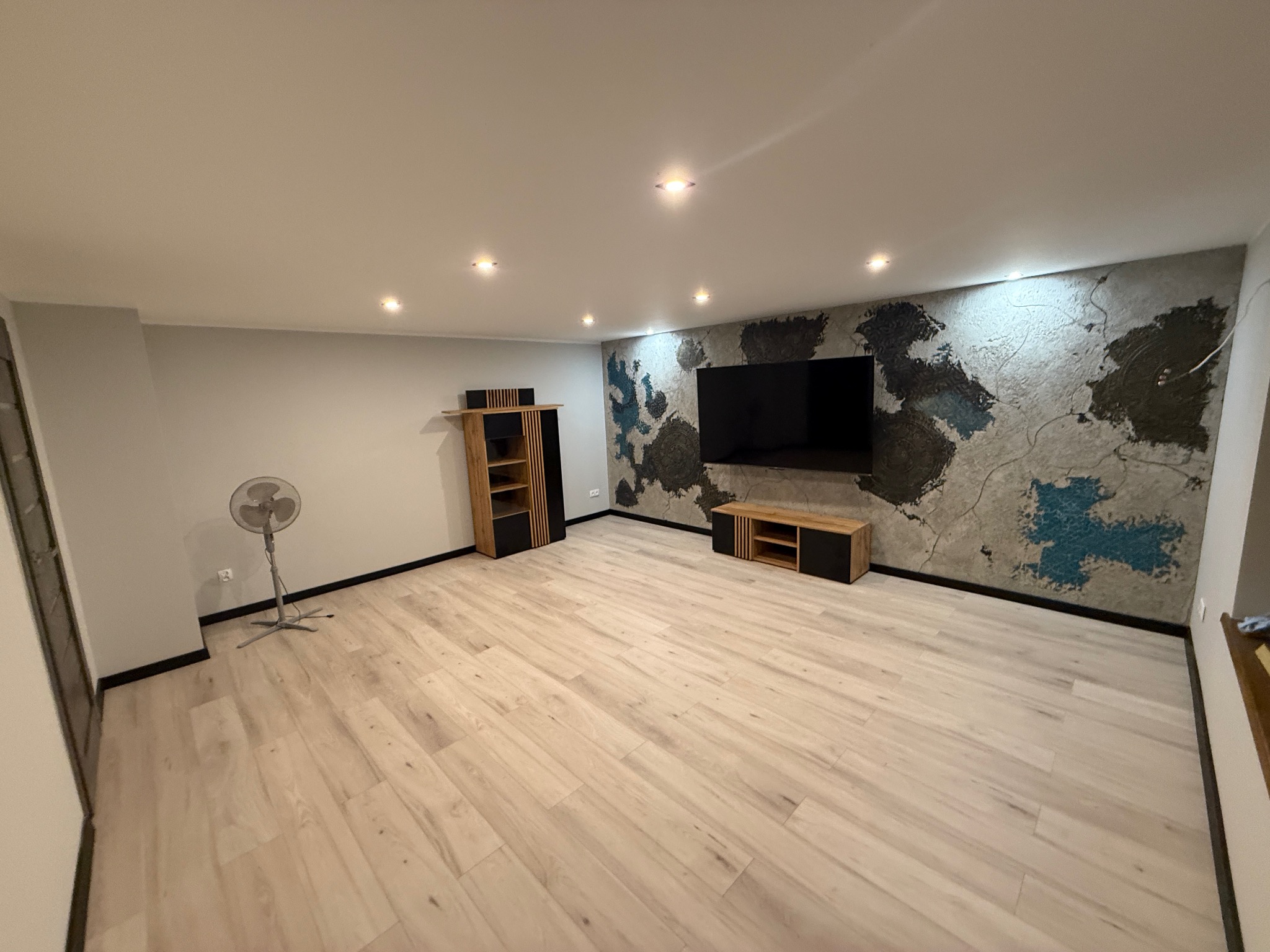 Jasne panele podłogowe w pokoju z dekoracyjną ścianą w stylu mapy świata, szafki RTV i wentylator. Oświetlenie sufitowe punktowe.