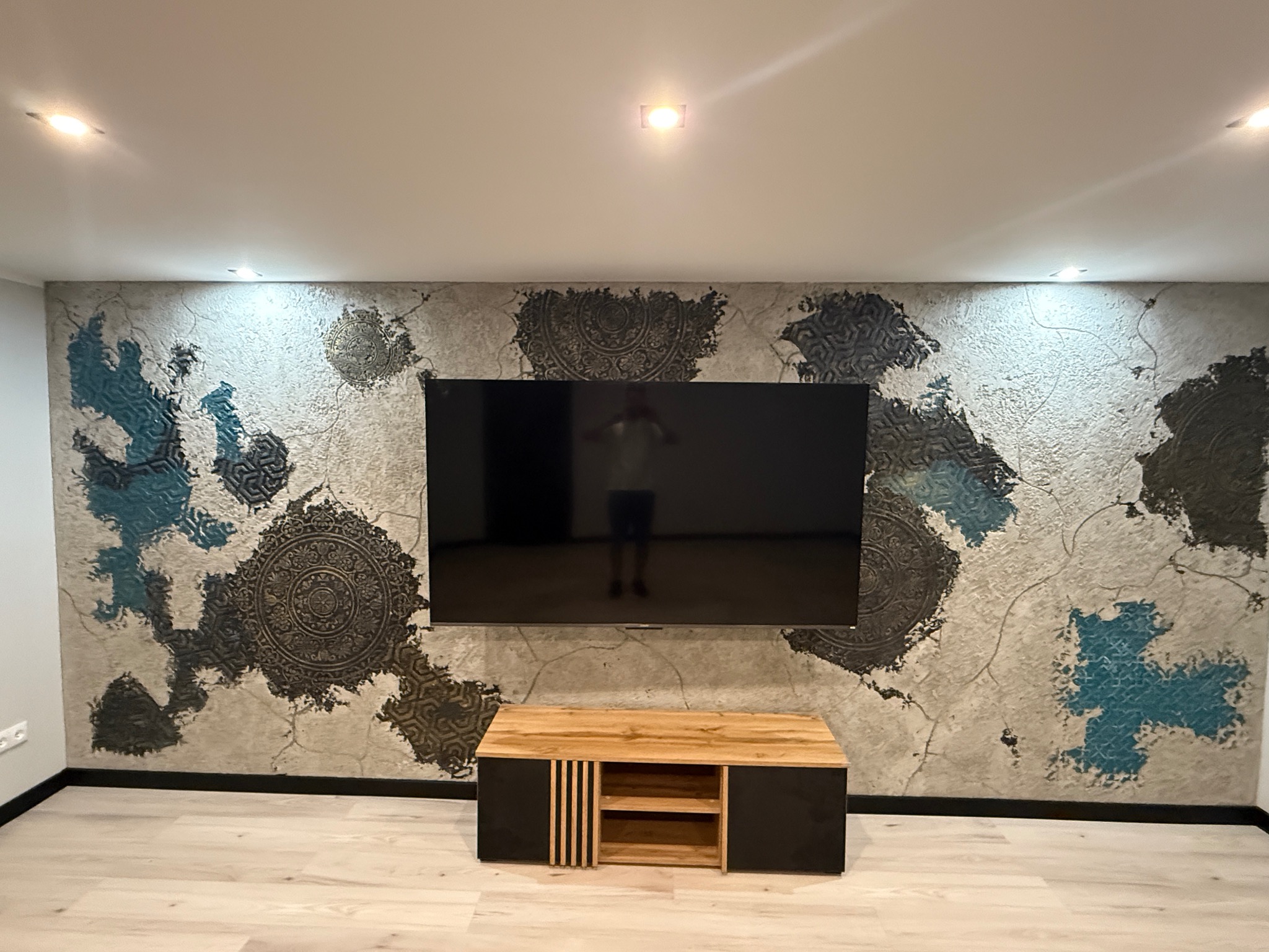 Ściana z dekoracyjnym tynkiem strukturalnym w odcieniach szarości i turkusu, z motywami mandali. Pod ścianą szafka RTV w kolorach czerni i drewna.