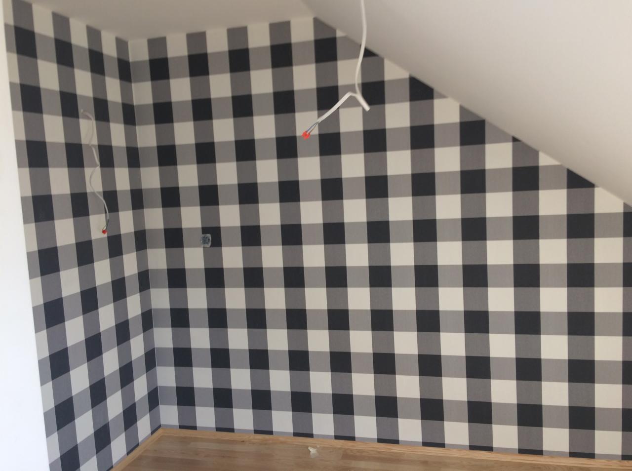 Ściana w pokoju na poddaszu pokryta tapetą w dużą czarno-białą kratę gingham, widoczne wystające kable elektryczne i drewniana podłoga.