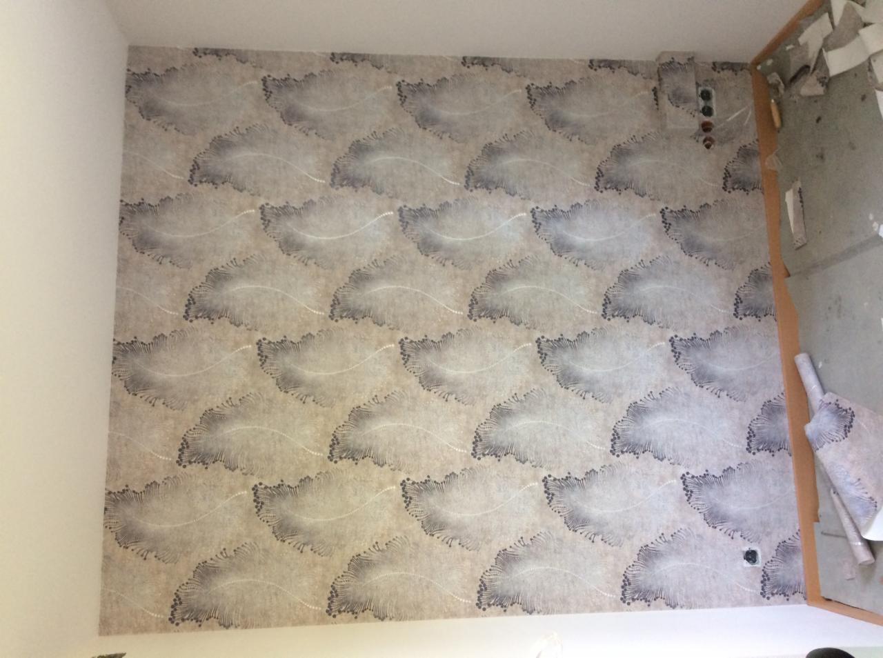 Ściana pokryta wzorzystą tapetą w odcieniach beżu i szarości z motywem przypominającym pióra, widoczne gniazdka elektryczne i fragment ściany w trakcie przygotowania do prac wykończeniowych.