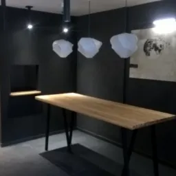 Wykończone wnętrze lokalu komercyjnego: czarne ściany, drewniane blaty stołów na metalowych nogach, dekoracyjne lampy wiszące i duży obraz z motywem gejszy.