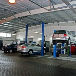 Wnętrze warsztatu samochodowego z trzema samochodami, jeden na podnośniku dwukolumnowym, drugi na czterokolumnowym, trzeci zaparkowany. Widoczna brama garażowa i oświetlenie sufitowe.