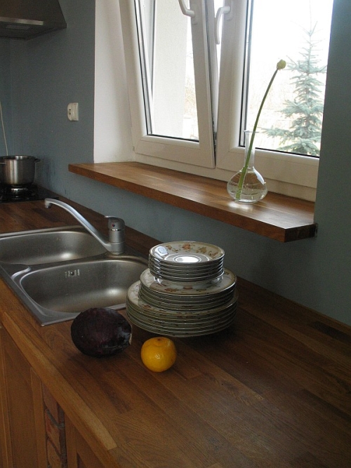 Blat dębowy w kuchni z umywalką, stosem talerzy, śliwką i pomarańczą, pod oknem z drewnianym parapetem, na którym stoi wazon z pojedynczym kwiatem.
