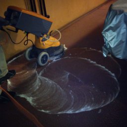 Komplex Styl Jan Bereś - Profesjonalna maszyna do czyszczenia podłóg w trakcie pracy, pozostawia ślady piany na brązowej powierzchni. Widoczny fragment osoby obsługującej urządzenie oraz elementy wyposażenia pomieszczenia.