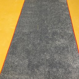 Komplex Styl Jan Bereś - Szary dywan z czerwonym obszyciem leży na żółtej podłodze. Widok z góry, równomierne oświetlenie. Dywan po czyszczeniu, gotowy do użytku.