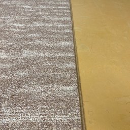 Komplex Styl Jan Bereś - Fragment podłogi z nową, wzorzystą wykładziną dywanową, przylegającą do starej, żółtej powierzchni. Widoczny brzeg łączenia materiałów.