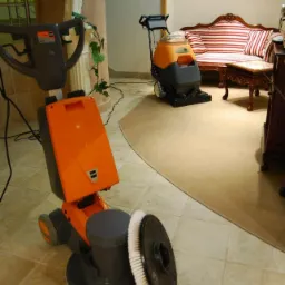 Dwie maszyny czyszczące na tle klasycznego wnętrza: jedna z pomarańczową obudową i szczotką, druga w odcieniach szarości i pomarańczy, obok sofa w paski i dekoracyjna kolumna z rzeźbą.