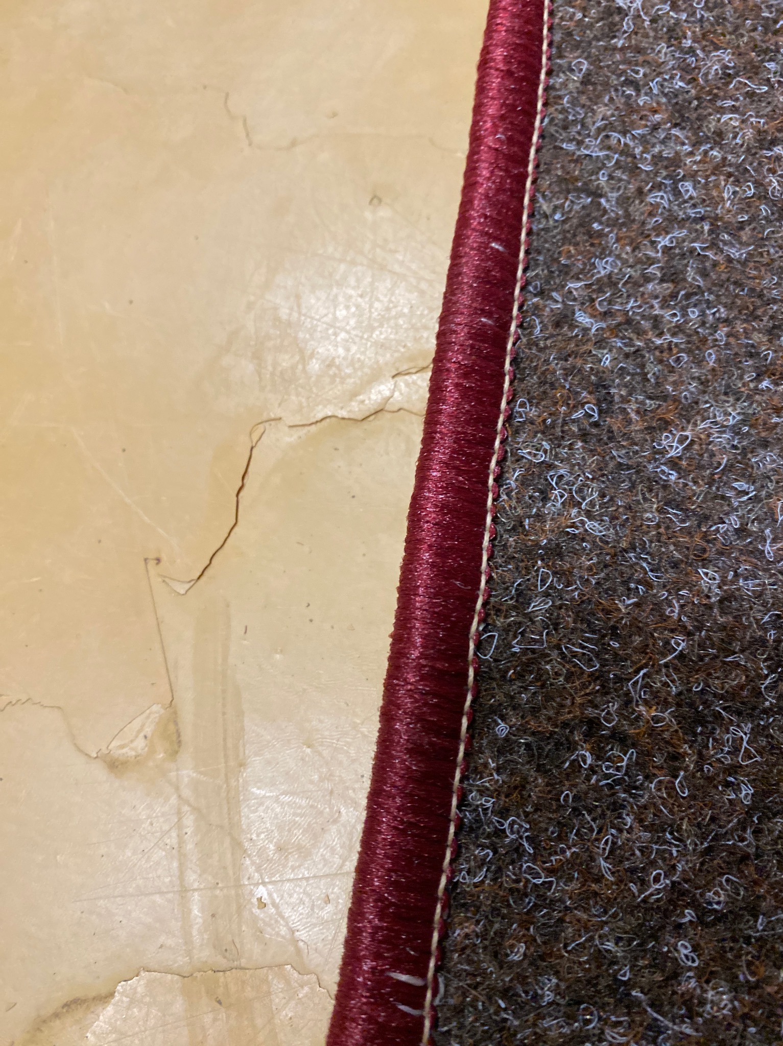 Fragment brązowego dywanu z bordową krawędzią na tle jasnej, zniszczonej podłogi. Widoczne detale struktury dywanu i uszkodzenia podłoża.