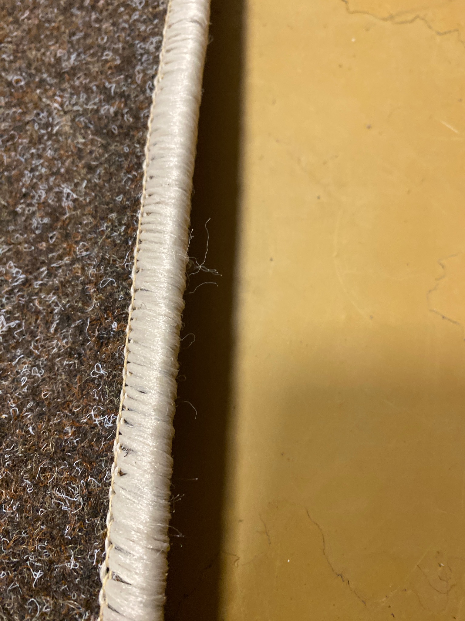 Brązowy dywan z jasnym obszyciem na żółtym tle. Widoczne włókna i struktura materiału w zbliżeniu. Detal wykończenia krawędzi dywanu.