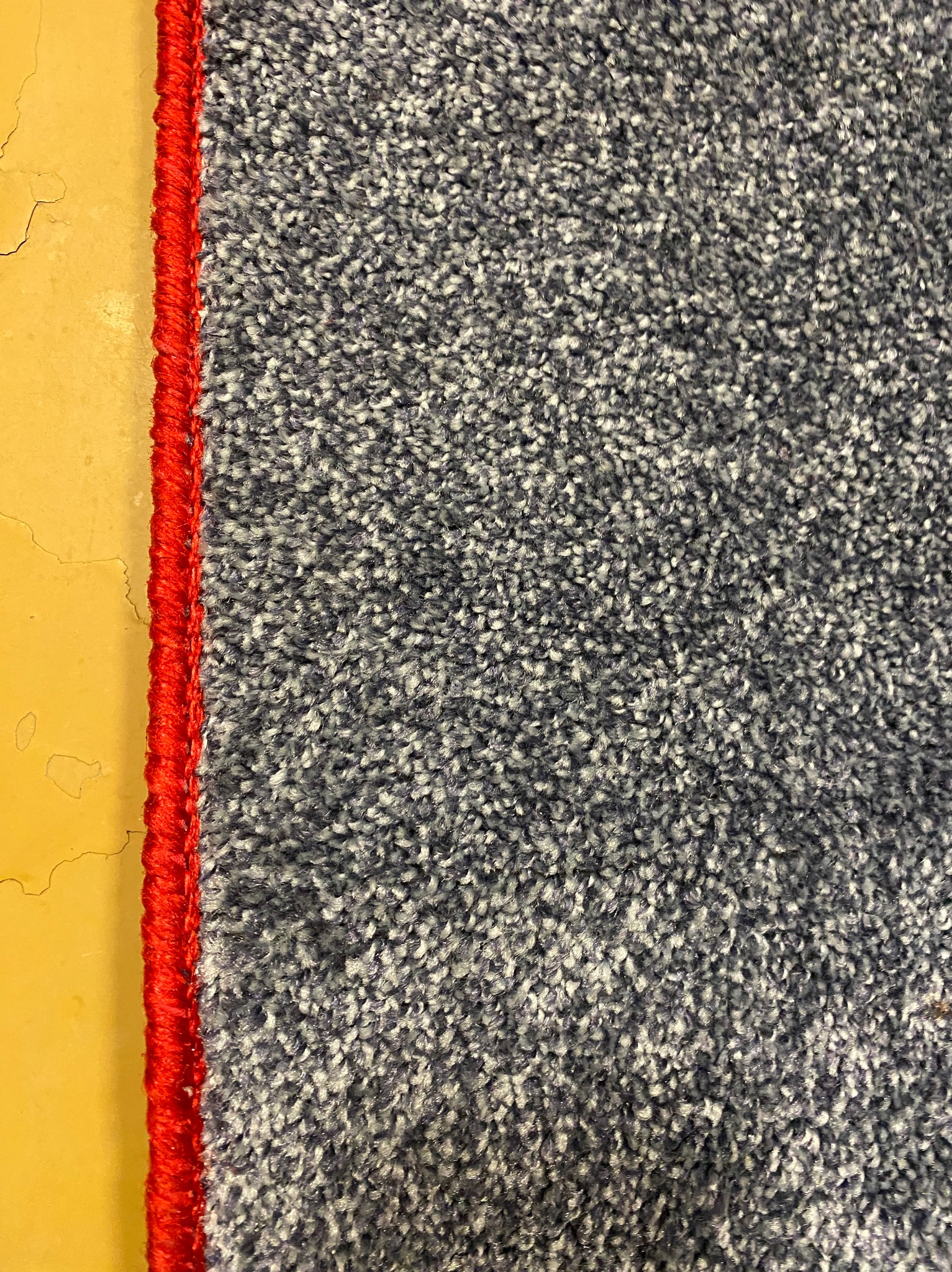 Szczegółowe zbliżenie na szaro-biały dywan z czerwonym obszyciem, leżący na żółtej podłodze z widocznymi pęknięciami. Tekstura dywanu jest wyraźnie widoczna.