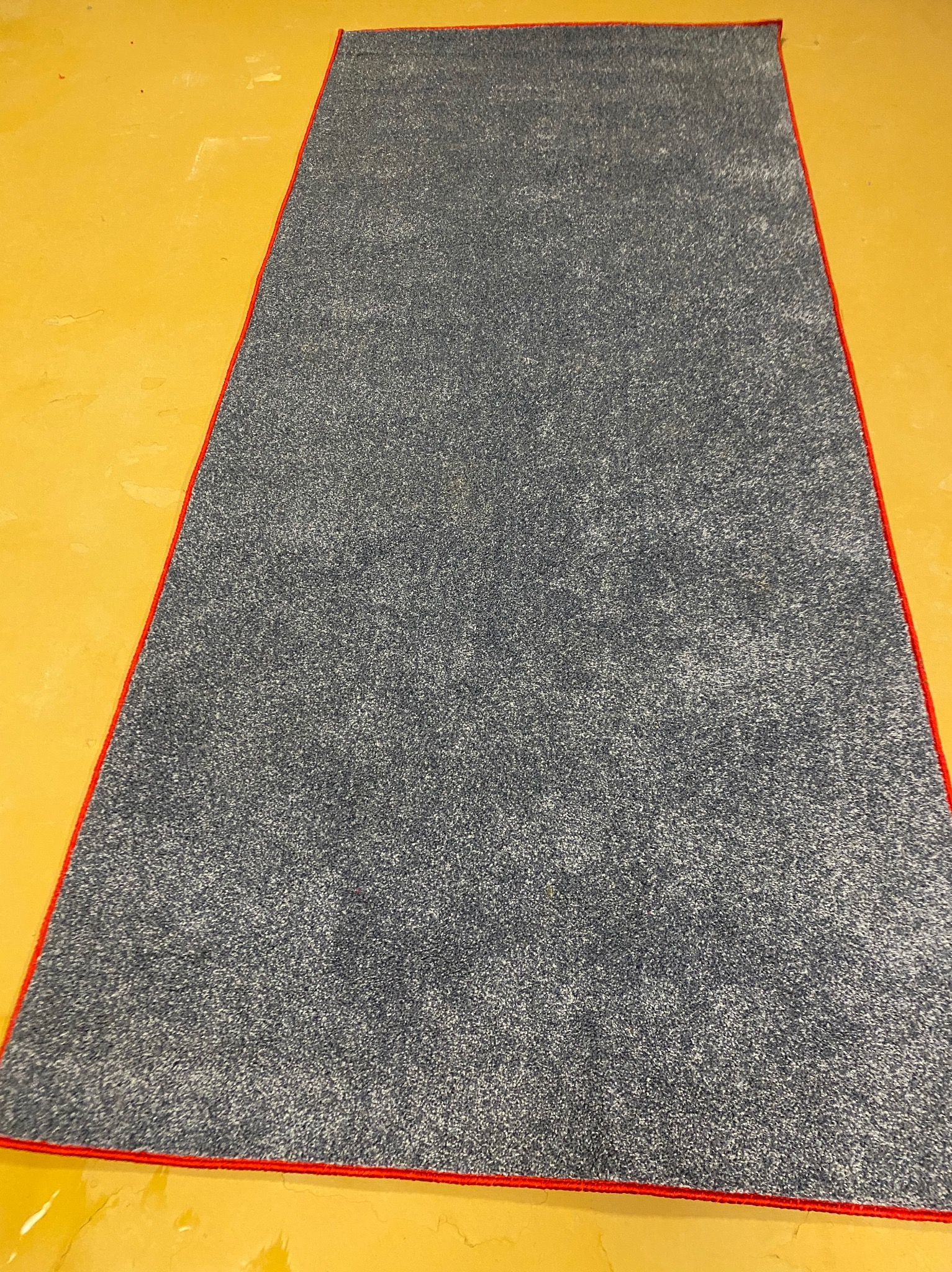 Szary dywan z czerwonym obszyciem leży na żółtej podłodze. Widok z góry, równomierne oświetlenie. Dywan po czyszczeniu, gotowy do użytku.