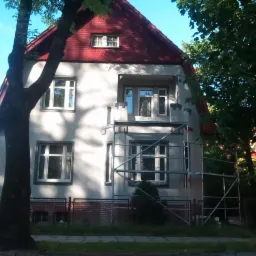 Dwupiętrowy dom z czerwoną dachówką, częściowo osłonięty rusztowaniem podczas prac elewacyjnych, z widocznymi drzewami w tle.