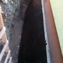 Wykop fundamentowy z zabezpieczoną, czarną powłoką hydroizolacyjną, widoczny fragment ściany budynku i drewniana drabina.