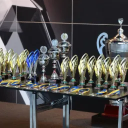 Rzędy złotych i srebrnych pucharów oraz medali z niebiesko-żółtymi wstążkami ustawione na czarnym stole z metalowymi nogami na tle czarnej ściany z abstrakcyjnym białym wzorem.