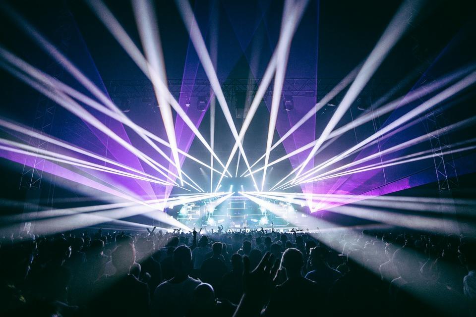 Tłum ludzi na koncercie oświetlony purpurowymi i białymi laserami, scena w tle.