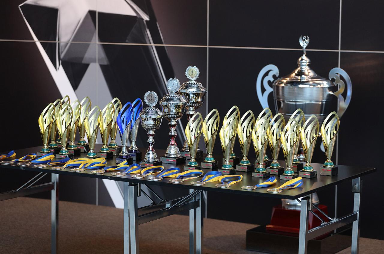 Rzędy złotych i srebrnych pucharów oraz medali z niebiesko-żółtymi wstążkami ustawione na czarnym stole z metalowymi nogami na tle czarnej ściany z abstrakcyjnym białym wzorem.