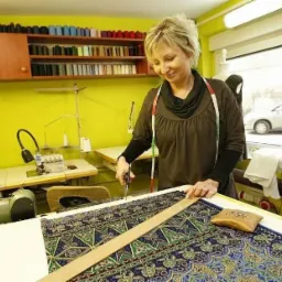 Kobieta z blond włosami w pracowni, przy stole krojąca wzorzystą tkaninę nożyczkami, z miarą na szyi, obok poduszeczka na igły, w tle maszyny do szycia i okno.