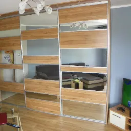 Duża szafa przesuwna z lustrami i drewnianymi panelami w salonie, odbicie sofy i telewizora w lustrach.