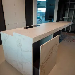 Wyspa kuchenna z blatem ze spieku kwarcowego o marmurkowym wzorze, z otwartą szafką i widocznym fragmentem płyty indukcyjnej. W tle okno i elementy wykończenia.